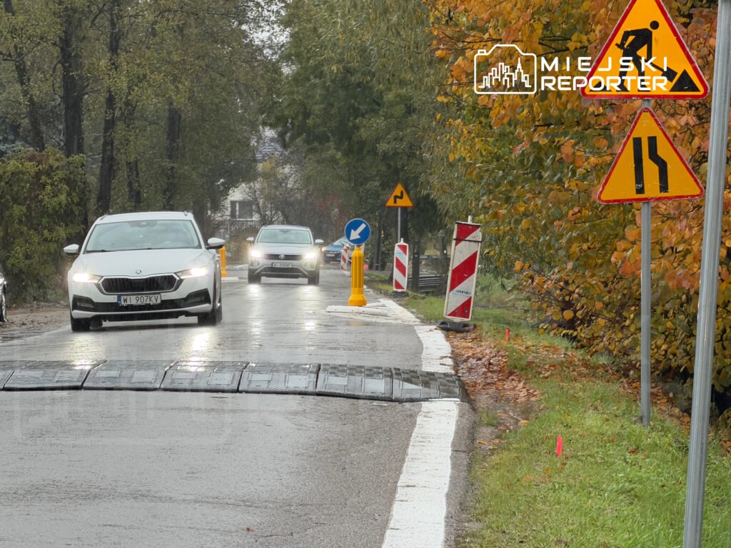 Kierowcy poruszają się po mokrej drodze z oznaczeniami drogowymi, w tle widoczne drzewa w jesiennych kolorach.