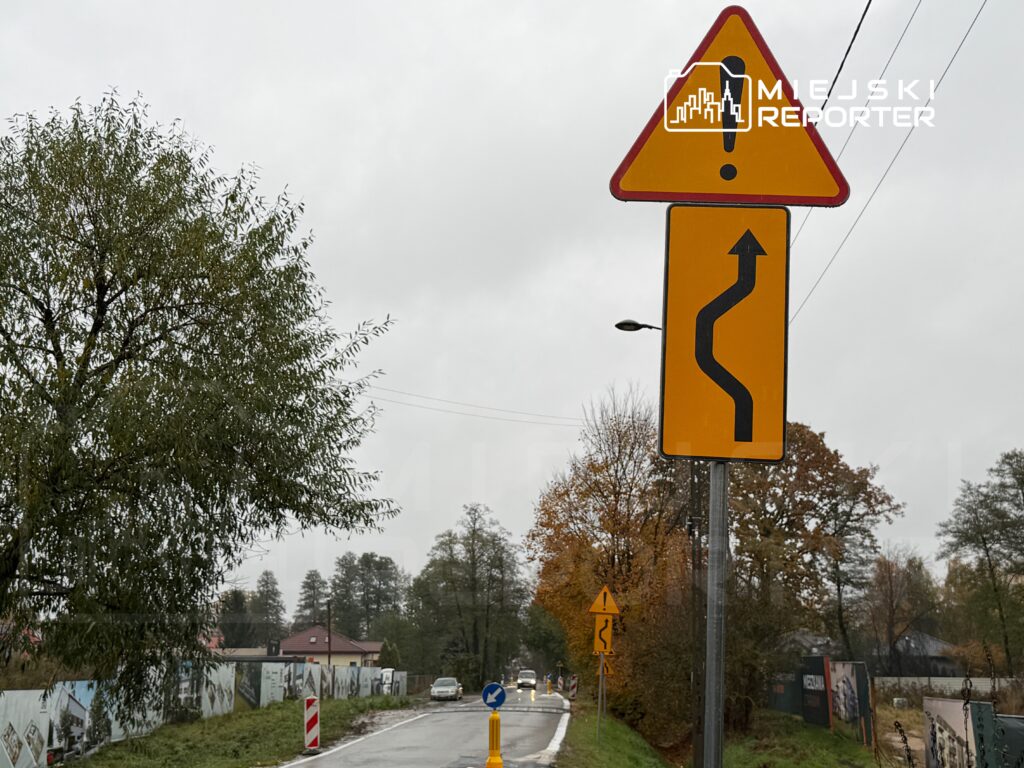 Droga z oznakowaniem informującym o zakrętach, otoczona drzewami i budynkami, w deszczowy dzień.