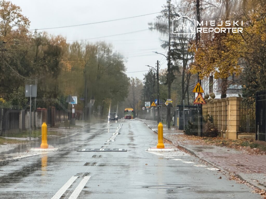 Deszczowa ulica z odbiciem na mokrym asfalcie, żółte słupki drogowe oraz oznakowanie drogowe w tle.