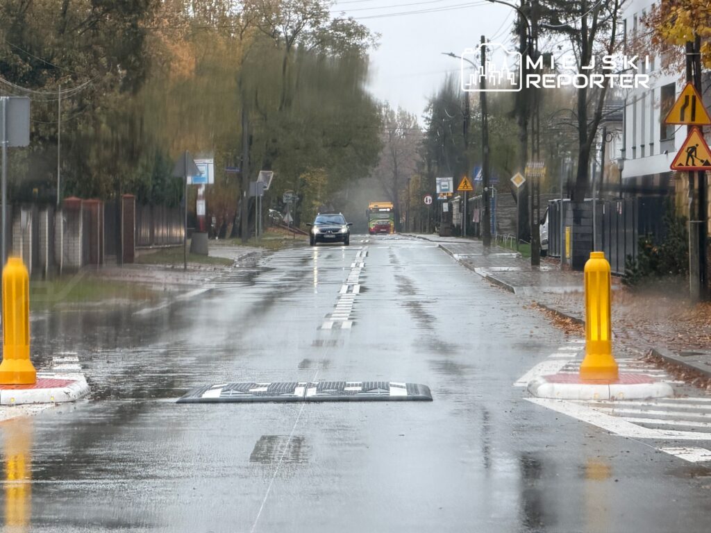 Ulica pokryta wodą po deszczu, z samochodem przejeżdżającym obok progu zwalniającego i znakami drogowymi w tle.