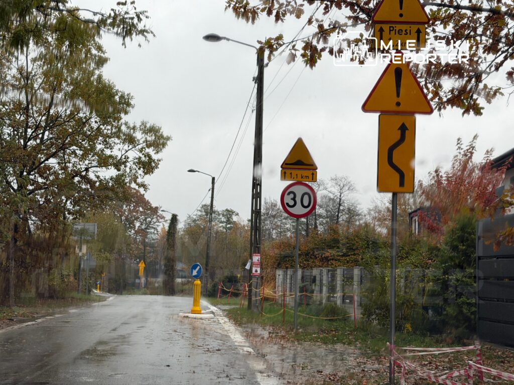 Droga w deszczowy dzień z widocznymi znakami drogowymi: ograniczenie prędkości do 30 km/h oraz ostrzeżenie o niebezpiecznym zakręcie.