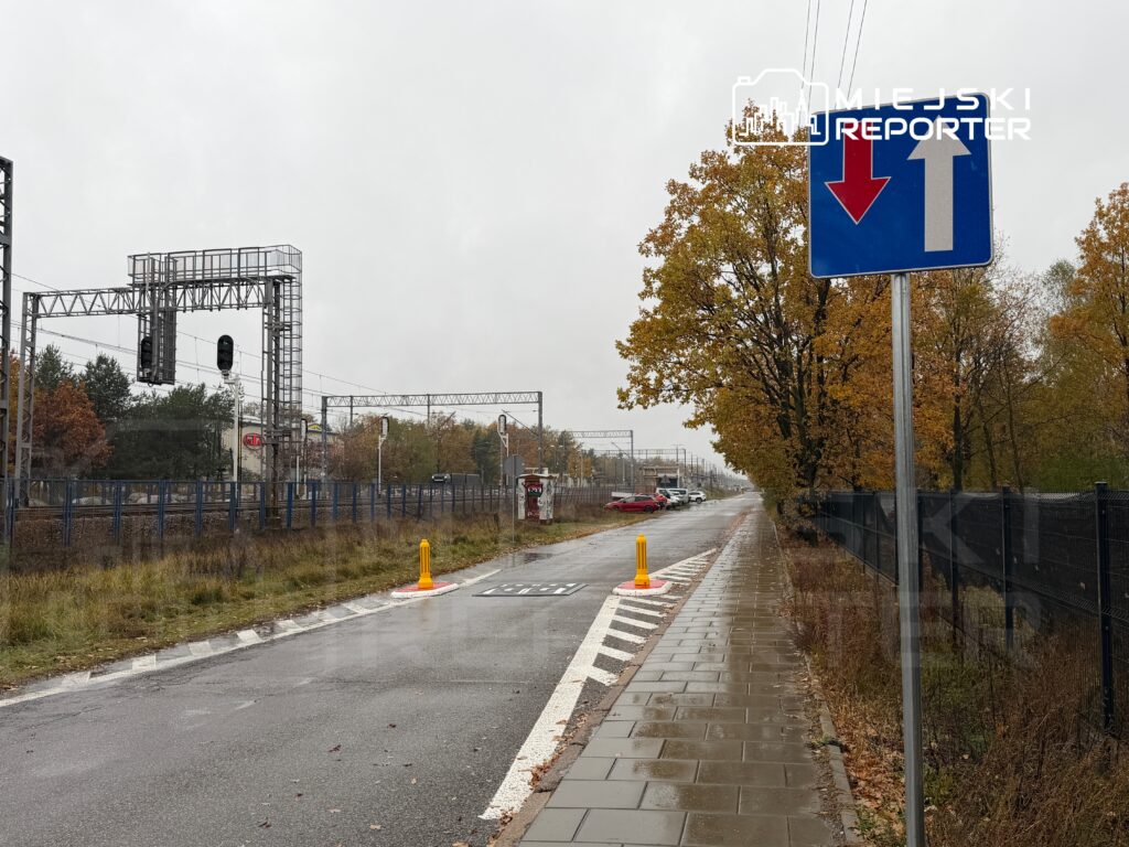 Droga z widokiem na tory kolejowe, znakiem drogowym wskazującym kierunek oraz drzewami w jesiennych barwach.