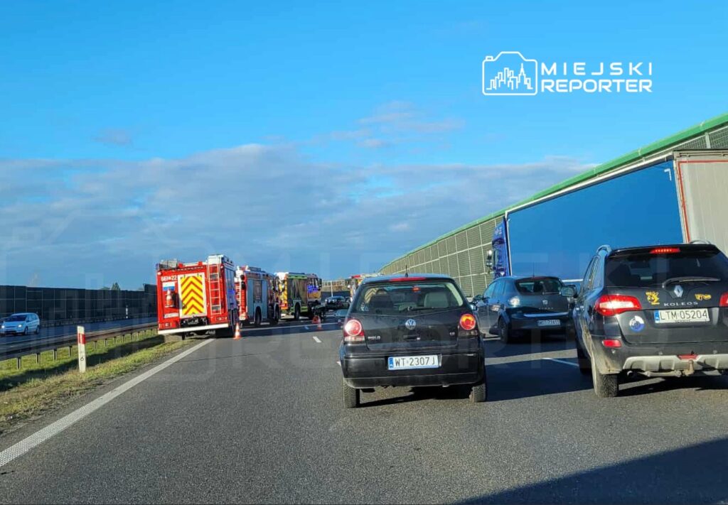 Na drodze zatrzymane samochody, w tle dwa wozy strażackie oraz służby ratunkowe ustawione przy barierkach.