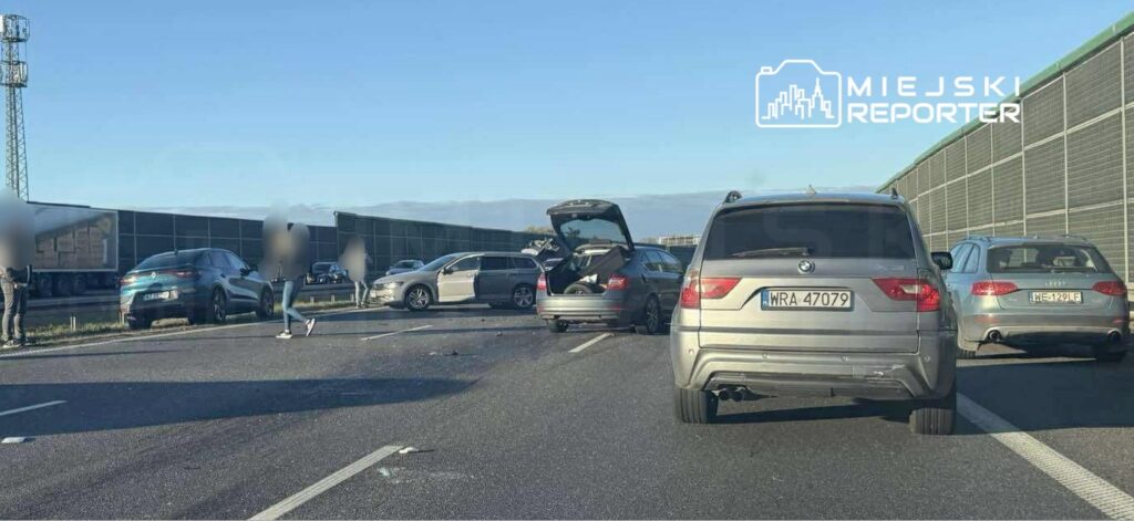 Wielu ludzi stoi obok uszkodzonych samochodów na autostradzie, w tle widać ciężarówki i ogrodzenie.