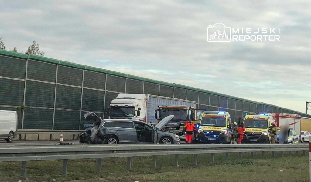 Na drodze, obok uszkodzonego samochodu osobowego, pracują ratownicy medyczni oraz strażacy, a w tle stoją ciężarówki.