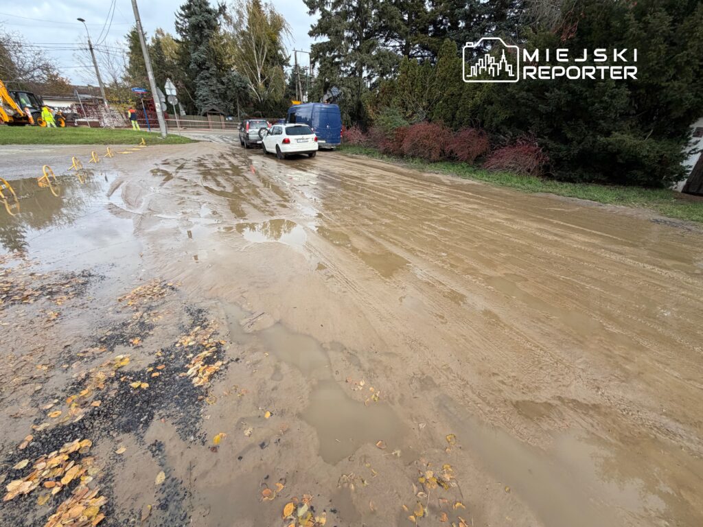 Na drodze widoczne kałuże i błoto, obok parkujące samochody oraz maszyny budowlane w tle.