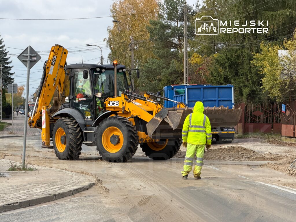 Maszyna budowlana JCB wykonuje prace ziemne na ulicy, obok pracownika w odblaskowej kamizelce.
