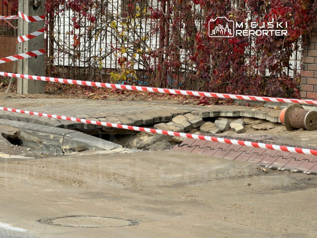 Uszkodzona nawierzchnia chodnika z widocznym zapadnięciem, ogrodzona taśmą ostrzegawczą. W tle zarośla i budynek.