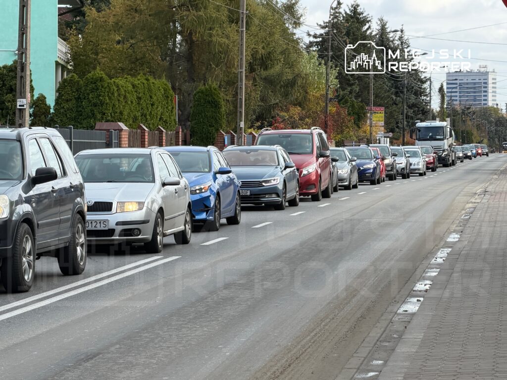 Ruch samochodowy wzdłuż ulicy z długim zatorze, w tle widać zieleń i budynki mieszkalne.