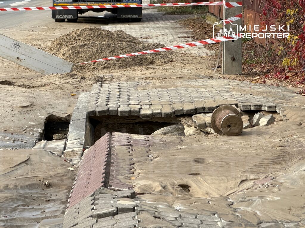 Uszkodzona nawierzchnia z kostki brukowej z odkrytym otworem oraz piaskiem, zabezpieczona taśmą ostrzegawczą.