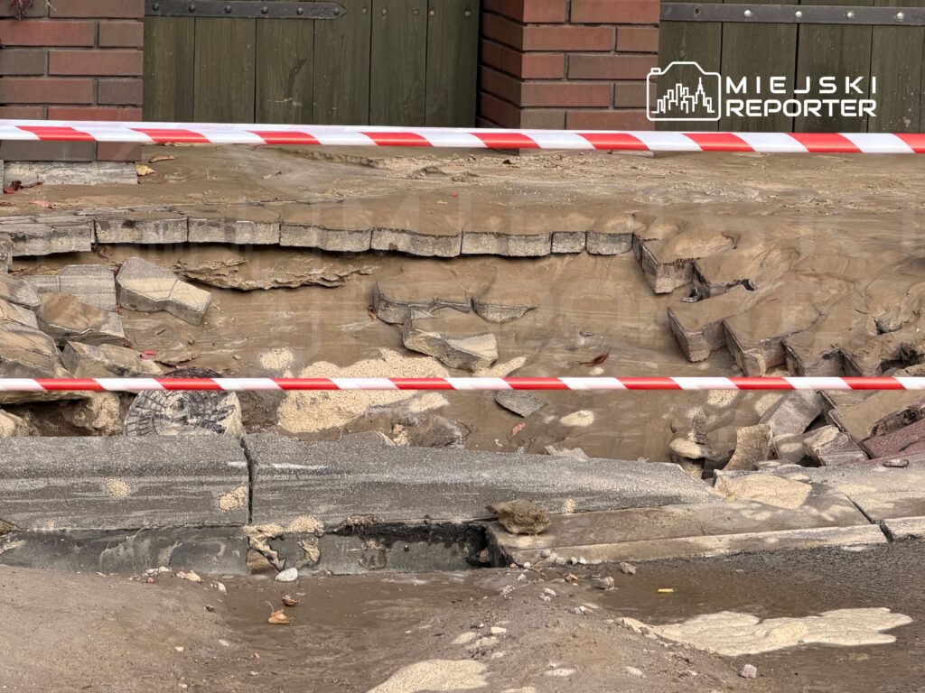 Uszkodzona nawierzchnia chodnika z widocznymi pęknięciami i odsłoniętą ziemią, zabezpieczona taśmą ostrzegawczą.