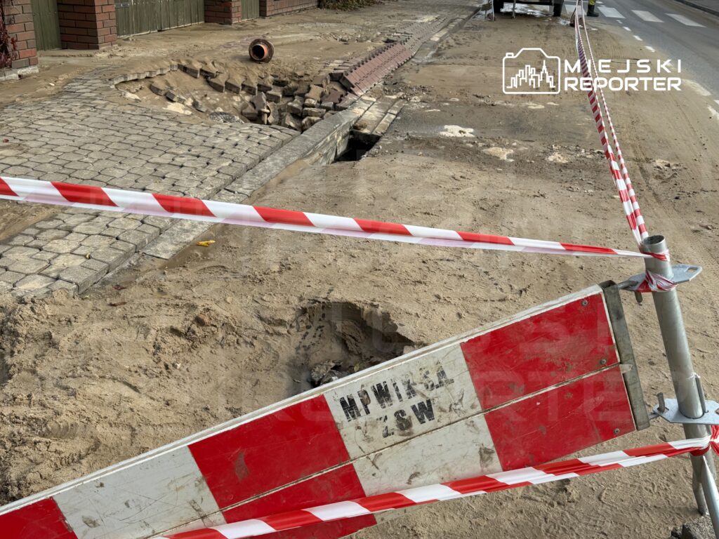 Usunięta kostka brukowa oraz wykop na drodze, otoczone barierkami z czerwono-białą taśmą ostrzegawczą.
