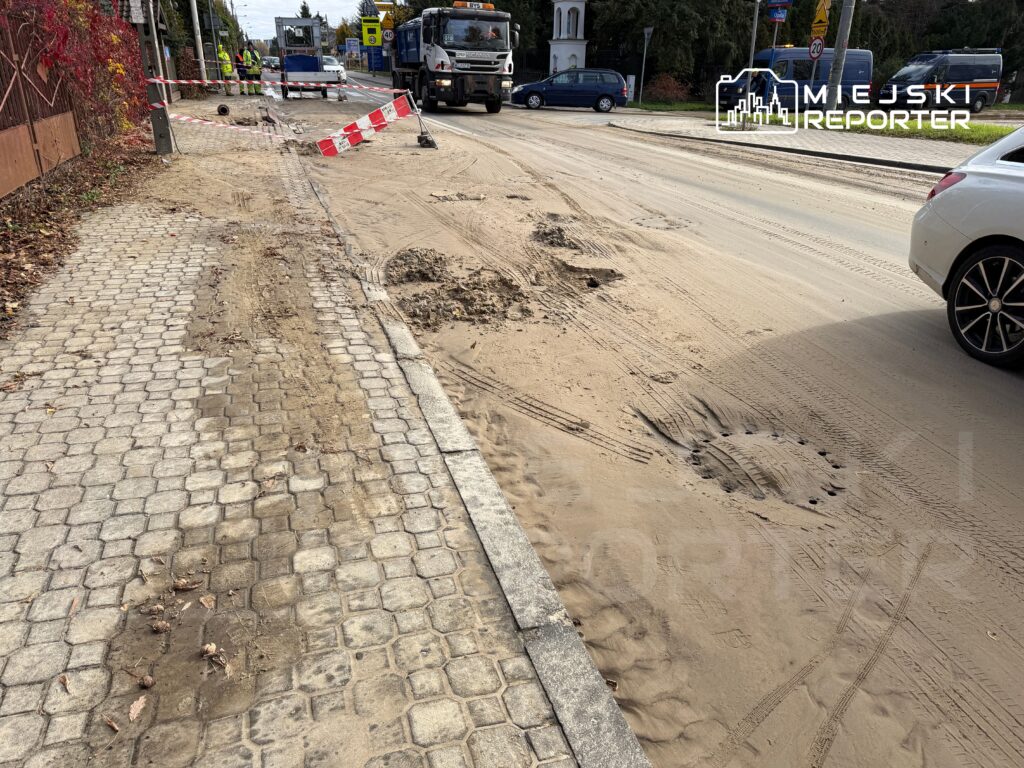 Robotnicy w odblaskowych kamizelkach pracują przy budowie drogi, wokół leżą resztki asfaltu i piasku.