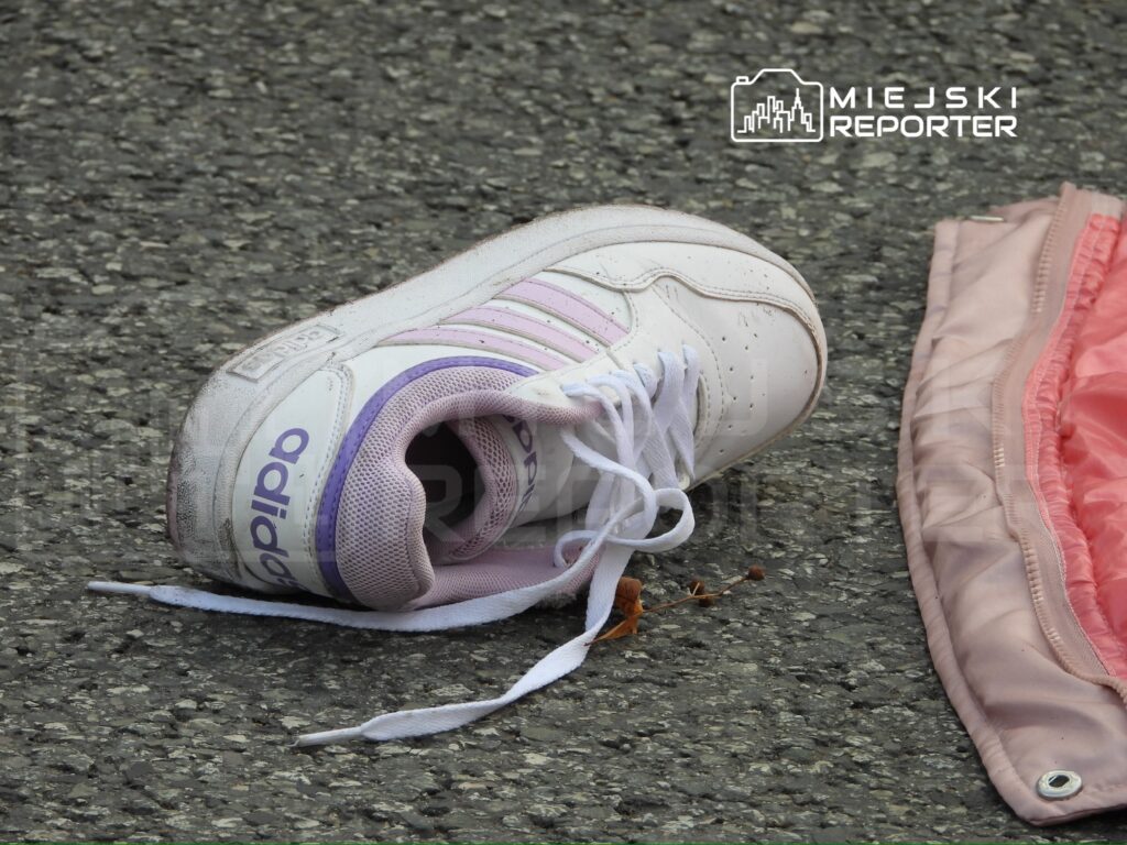Biały but sportowy z fioletowymi akcentami leży na asfaltowej nawierzchni obok różowego materiału.