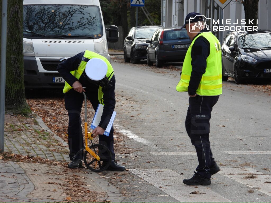 Policjant w kamizelce odblaskowej mierzy odległość za pomocą dalmierza, a drugi policjant obserwuje sytuację na ulicy.