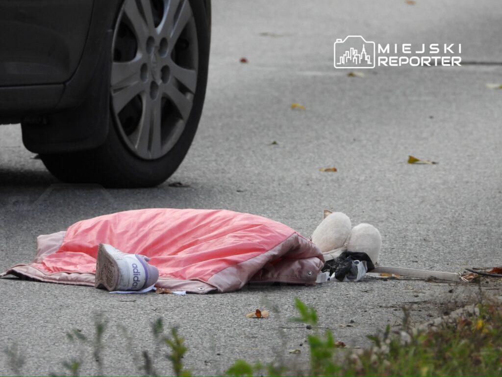 Na jezdni leży różowa kurtka obok białego buta sportowego, w tle widać koło samochodu.