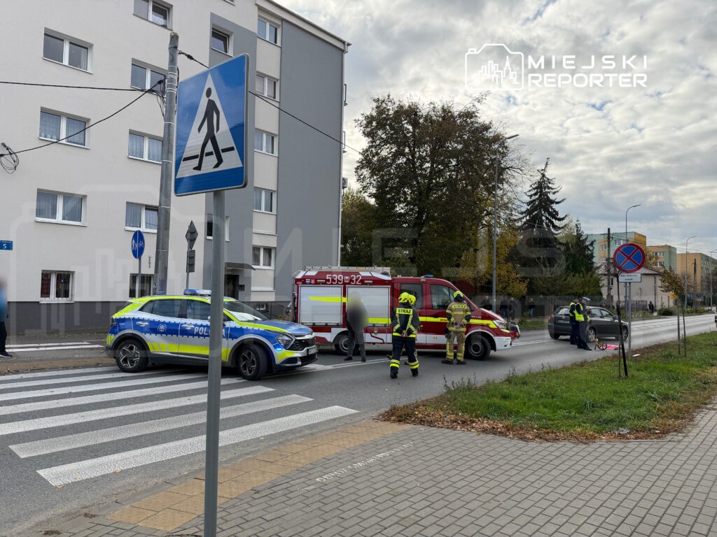 Na przejściu dla pieszych interweniują strażacy oraz policja, obok uszkodzony samochód. W tle widoczny budynek mieszkalny.