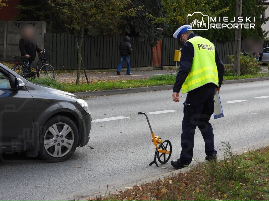 Policjant w kamizelce odblaskowej mierzy drogę przy zaparkowanym samochodzie na ulicy.