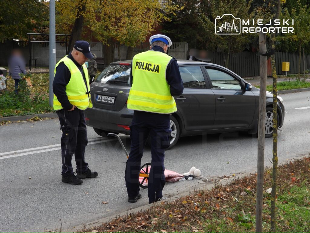 Policjanci w żółtych kamizelkach analizują miejsce wypadku drogowego obok zaparkowanego samochodu, na drodze leży uszkodzona zabawka.
