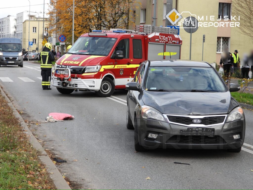 Strażacy w kamizelkach odblaskowych interweniują przy wypadku drogowym, obok leży różowa odzież na jezdni.