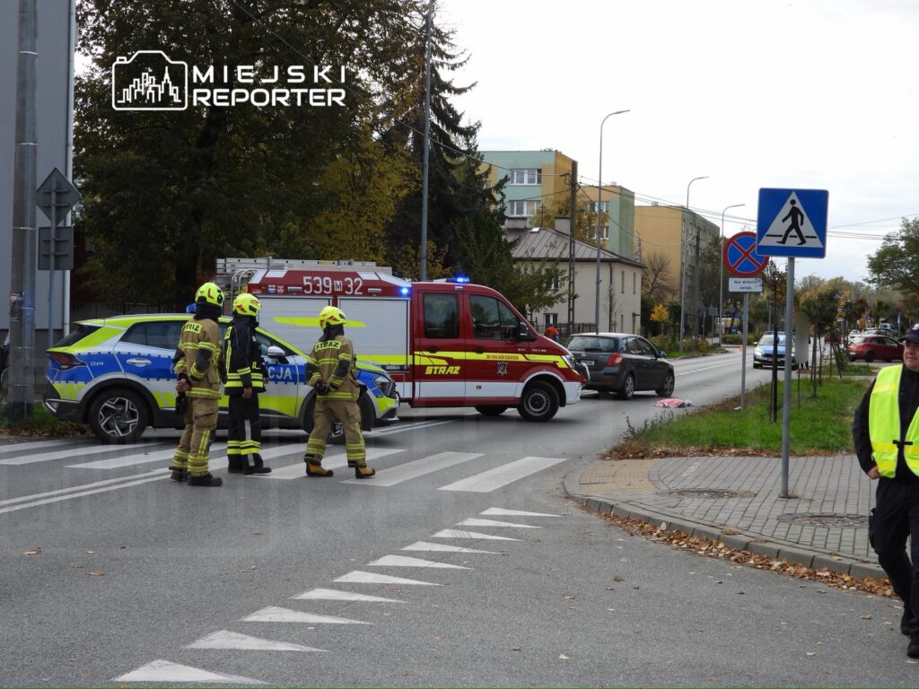 Czterech strażaków w kombinezonach ochronnych stoi przy wozie strażackim na skrzyżowaniu, obok karetki i oznakowania drogowego.