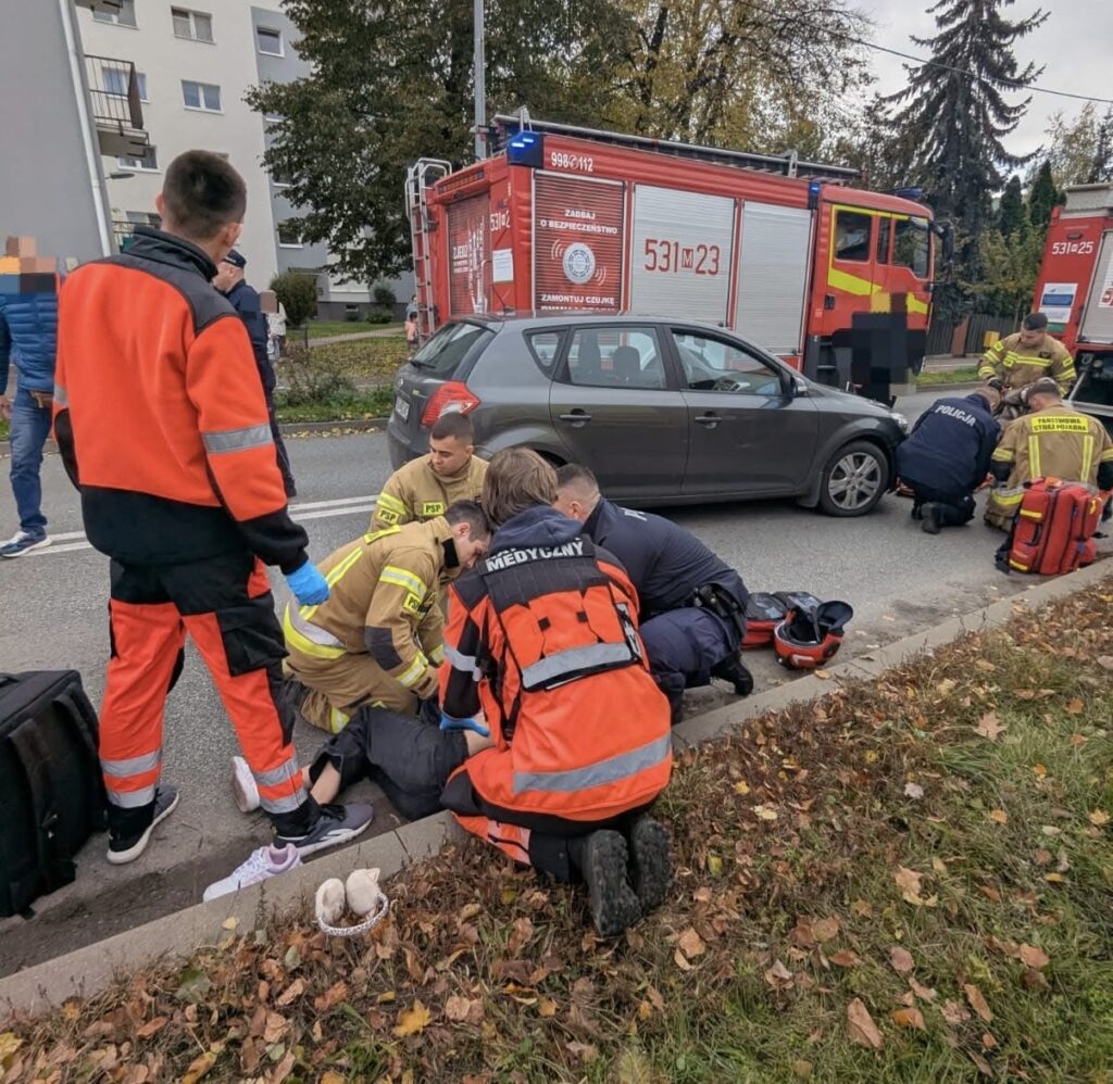 Grupa ratowników medycznych i strażaków udziela pomocy poszkodowanej osobie na chodniku obok drogi.