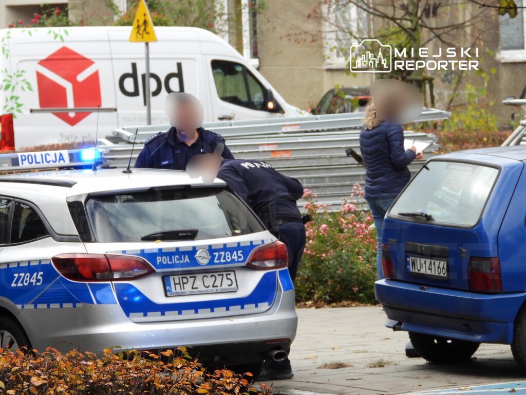 Policjanci w mundurach prowadzą działania przy radiowozie na miejskiej ulicy, obok stoi kobieta z notatnikiem.