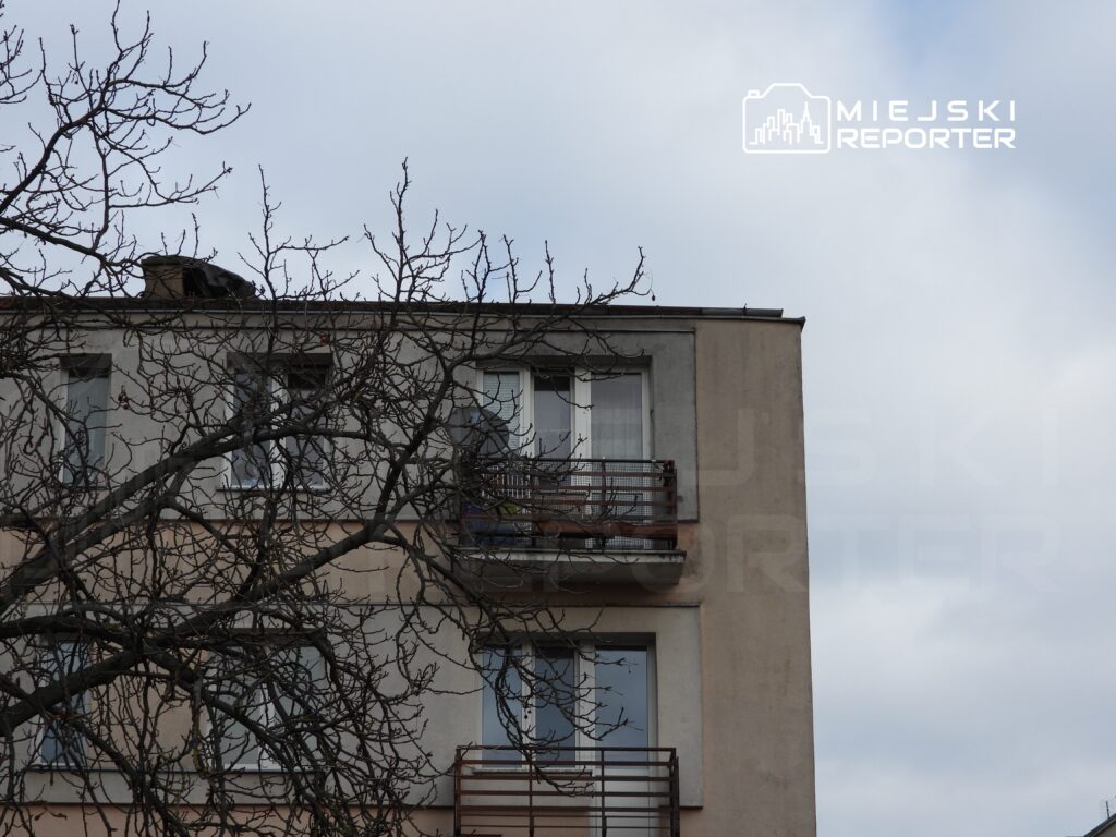 Czteropiętrowy blok mieszkalny z balkonem, w tle gołe gałęzie drzewa i pochmurne niebo.