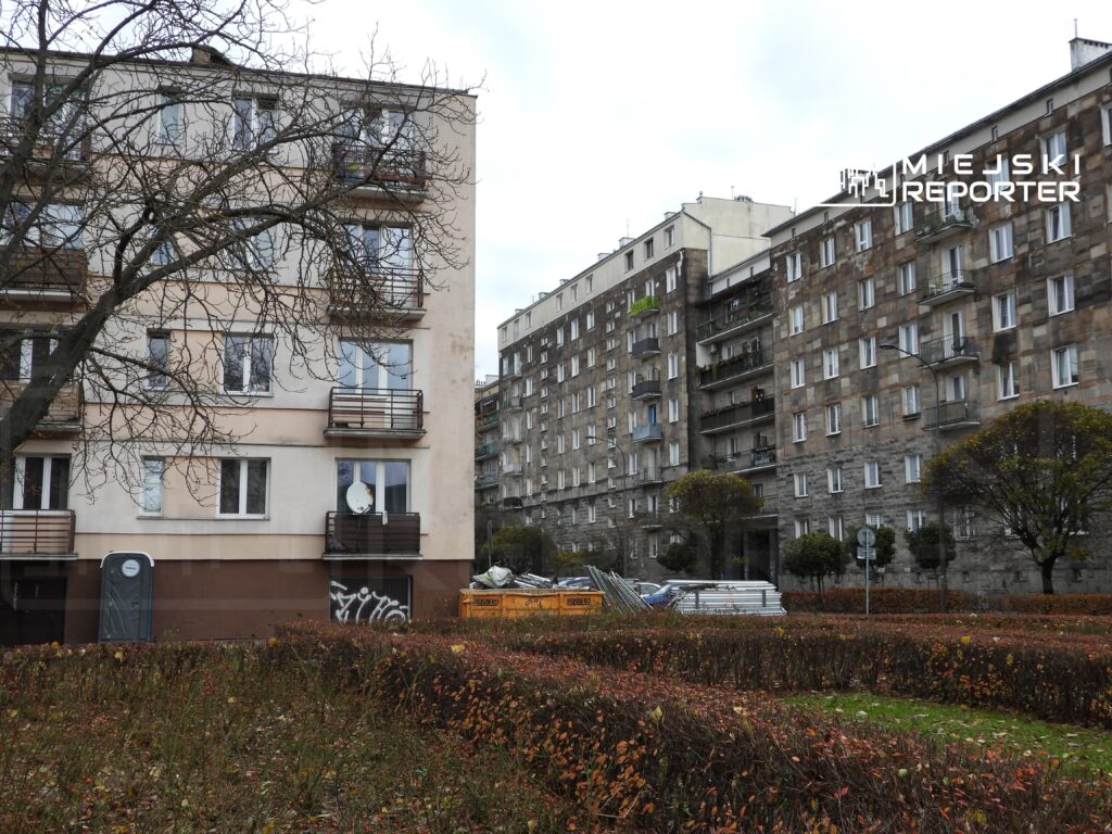 Nowoczesny budynek mieszkalny z balkonami oraz stary blok w tle, otoczone krzewami i drzewami w parku.