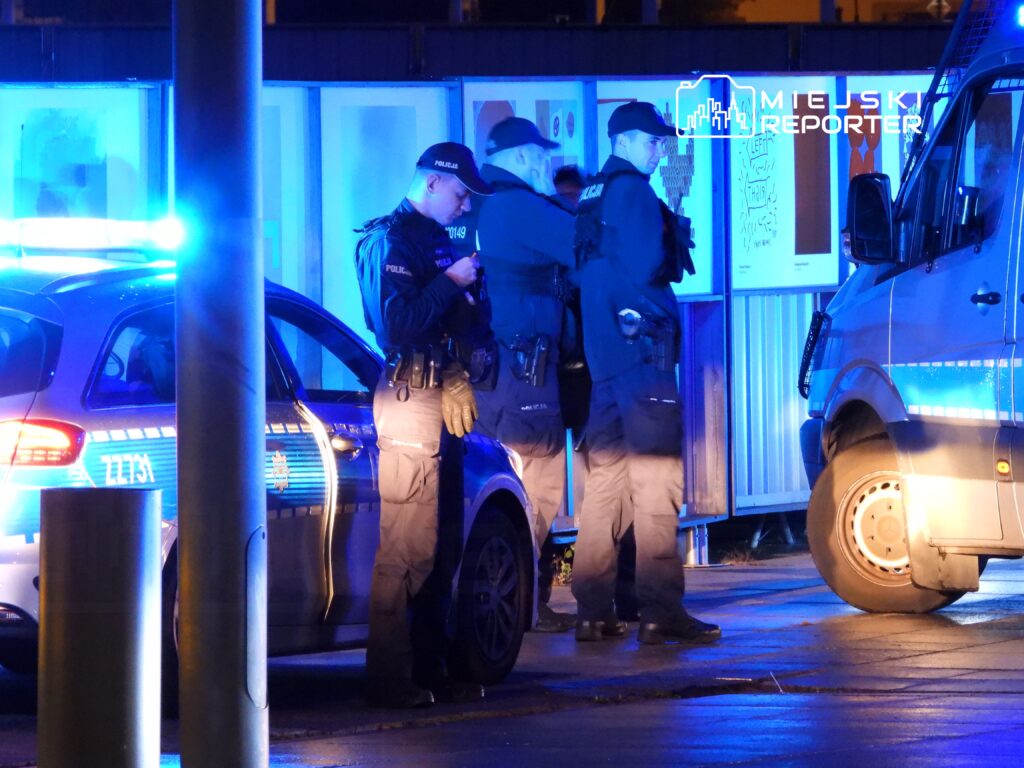 Grupa policjantów w mundurach stoi obok radiowozu z włączonymi sygnałami świetlnymi w nocnej scenerii.