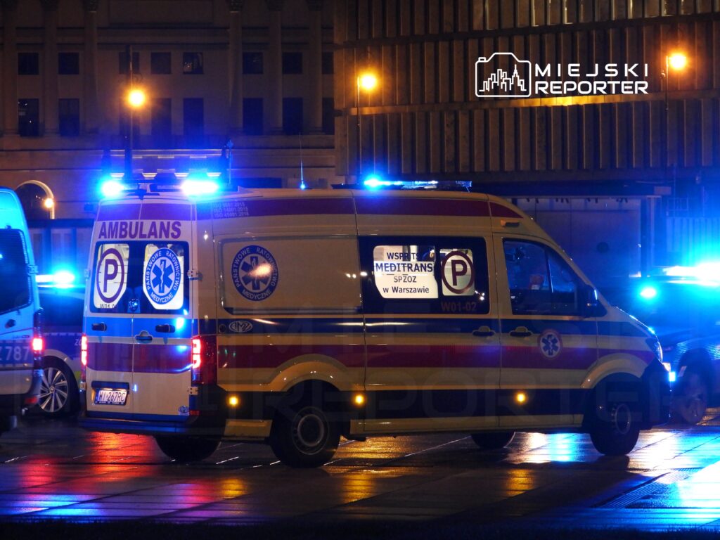 Karetka pogotowia z włączonymi sygnałami świetlnymi na nocnej ulicy, odbijająca światło na mokrej nawierzchni.
