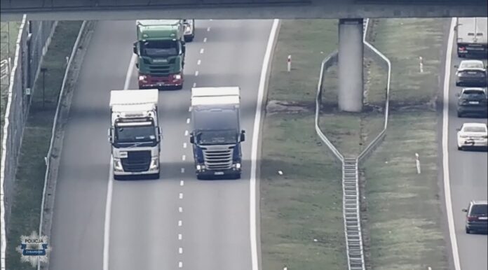 Trzy ciężarówki poruszają się po ruchliwej drodze, obok pasażerskie samochody i ogrodzenie w tle.