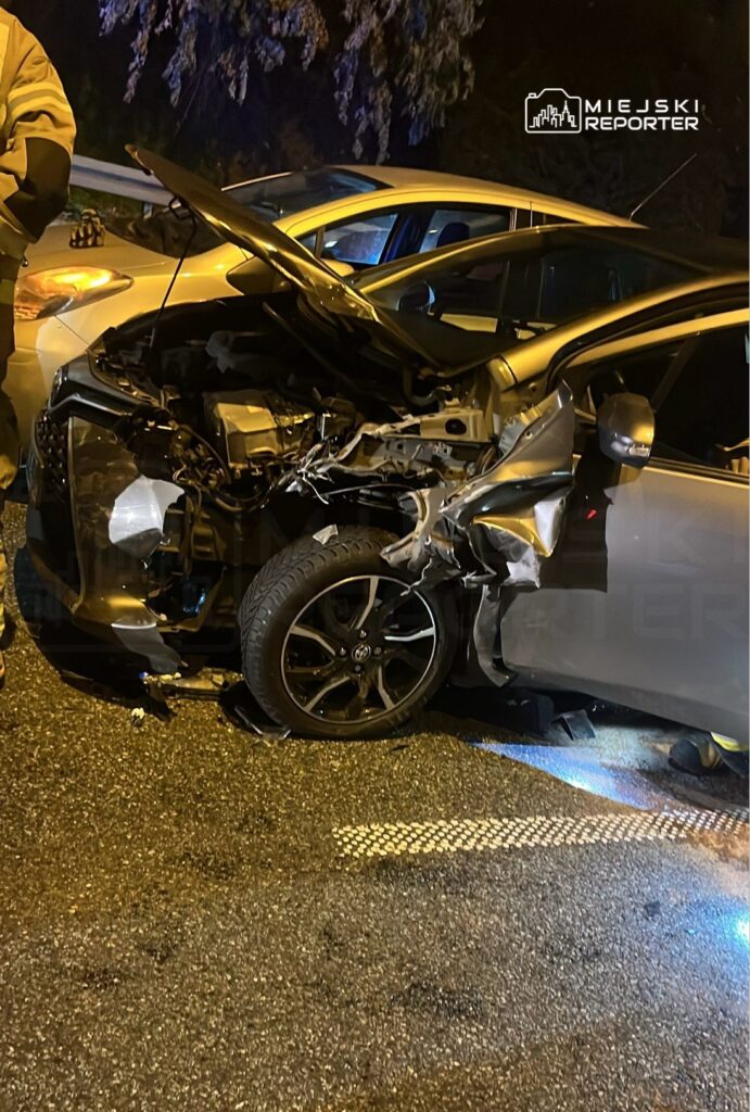 Uszkodzony samochód osobowy z rozdartą karoserią stoi na drodze, obok niego mężczyzna w mundurze.