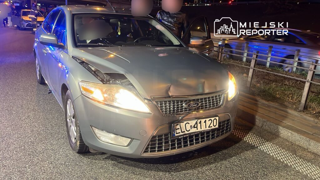 Ford z uszkodzoną maską stoi na drodze, otoczony przez policjantów i inne pojazdy w tle.