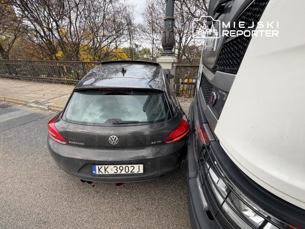 Czarny samochód osobowy Volkswagen Scirocco stłuczony przez dużą ciężarówkę na ulicy, w tle widoczna żeliwna balustrada.