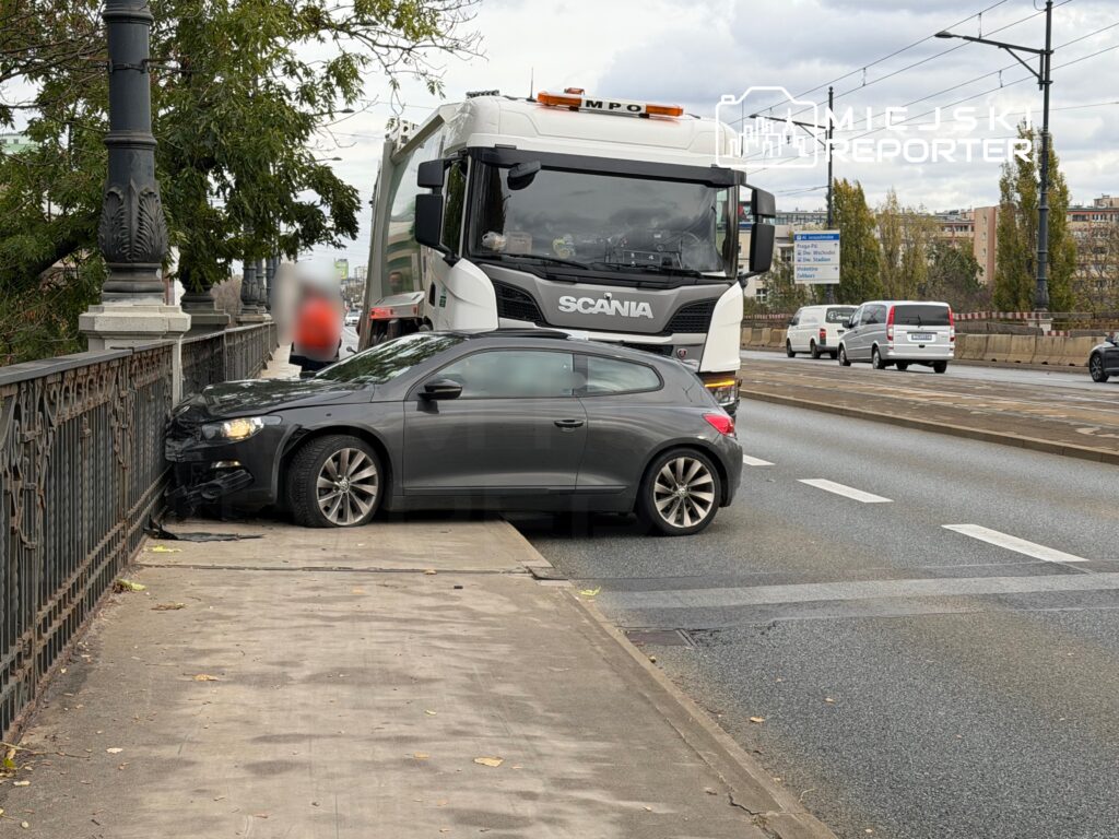 Czarny samochód osobowy stoi na chodniku, zderzony z barierką, obok niego przejeżdża ciężarówka.