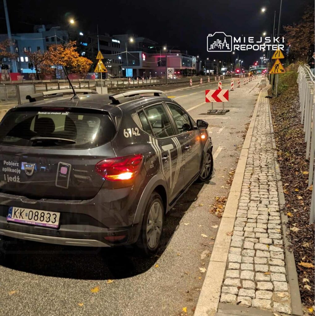 Samochód osobowy zaparkowany na ulicy nocą, obok oznakowania robót drogowych i barierek ochronnych.