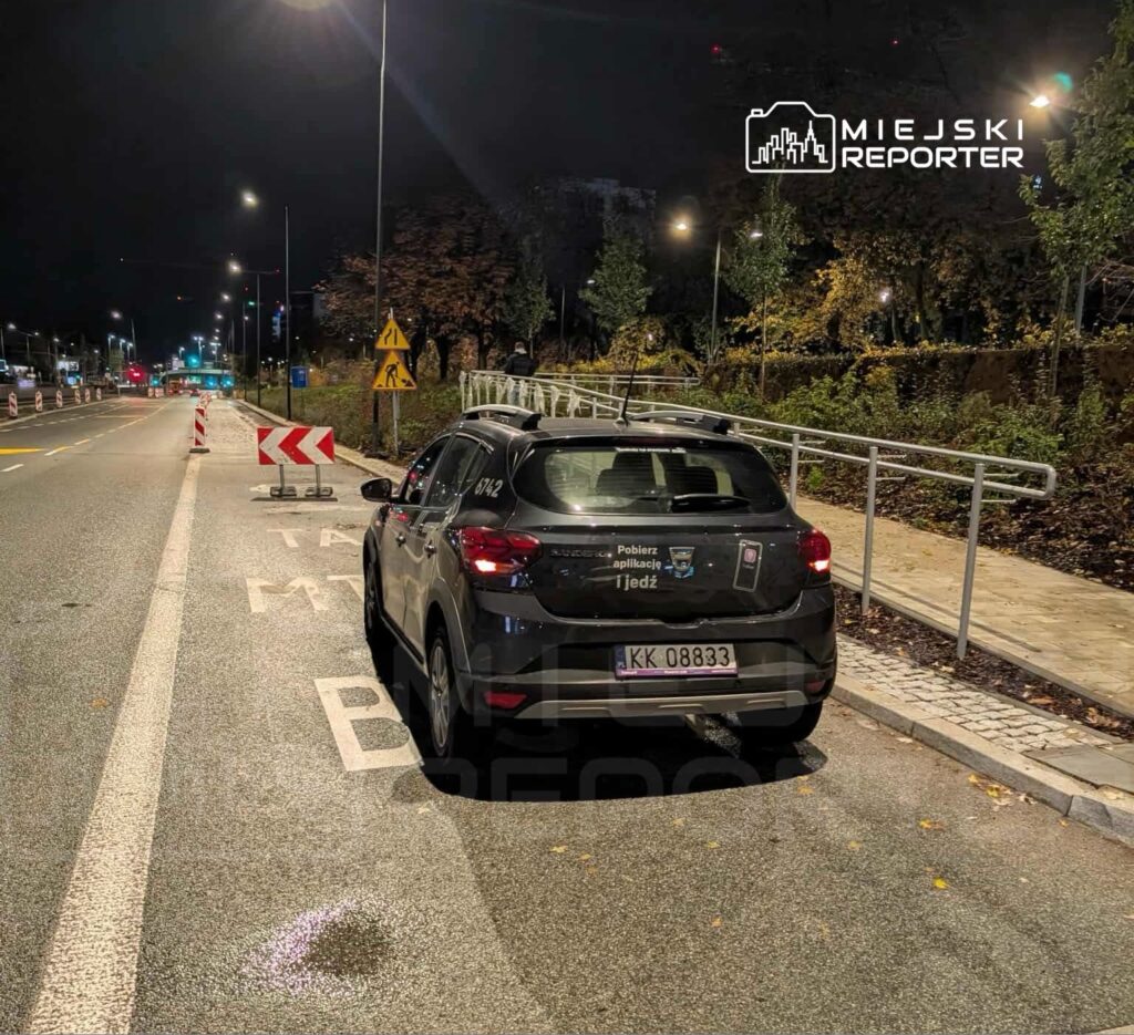 Czarny samochód zaparkowany na oświetlonej ulicy, obok oznakowanie robót drogowych oraz barierki ochronne.