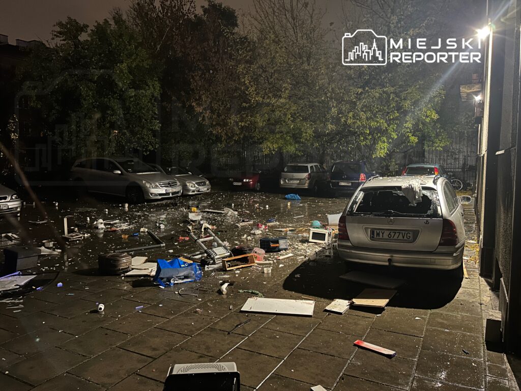 Rozrzucone śmieci i zniszczone przedmioty na parkingu, w tle zaparkowane samochody.