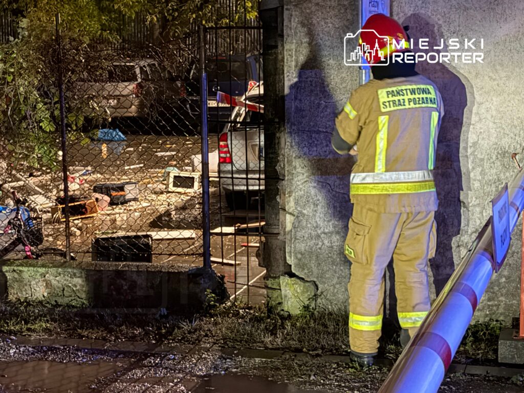 Strażak w mundurze z odblaskowymi elementami bada teren w zagraconym podwórku, w tle widać pojazdy i odpady.