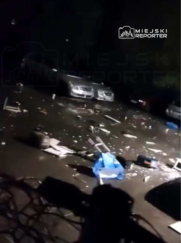 Ziemia pokryta odpadkami i gruzem, w tle dwa zaparkowane samochody. Scena nocna z chaotycznym otoczeniem.