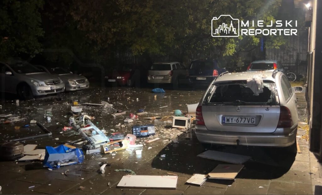 Zniszczony samochód osobowy na parkingu, wokół porozrzucane odpady i fragmenty sprzętu elektronicznego.