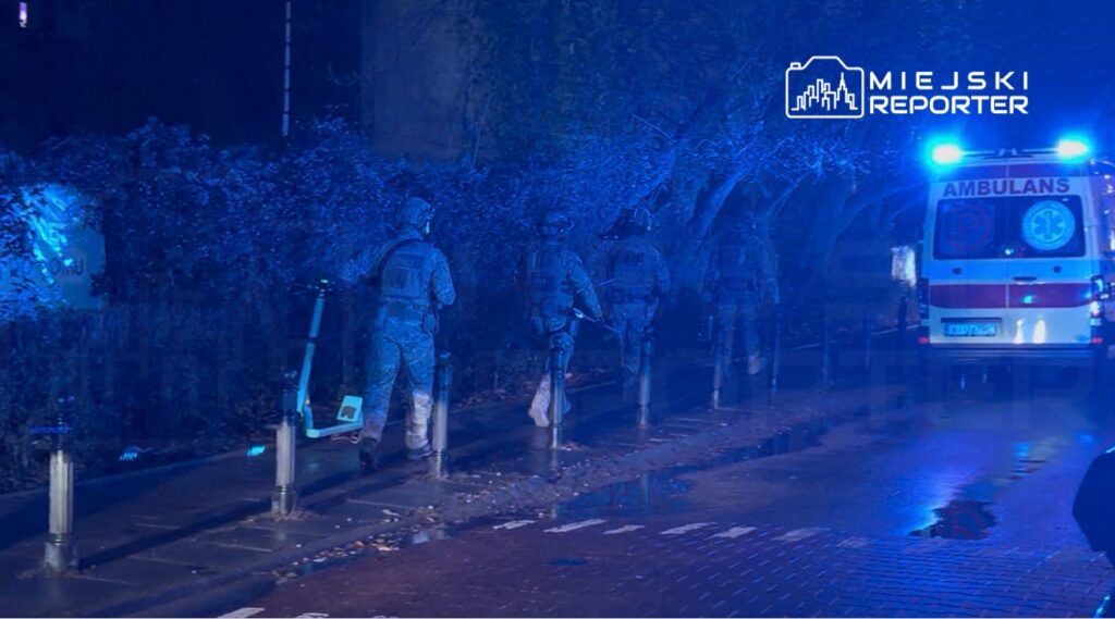 Grupa uzbrojonych żołnierzy patroluje oświetloną niebieskim światłem ulicę obok karetki pogotowia.