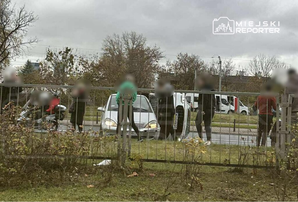 Grupa osób obserwuje przewrócone auto przy drodze, na tle szarego nieba i drzew.