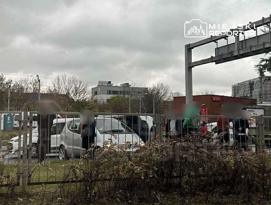 Grupa osób wokół zderzonego samochodu osobowego na miejskiej ulicy w pochmurny dzień.