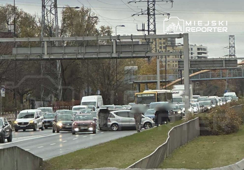 Ruch samochodowy na zatłoczonej drodze, z kilkoma pojazdami zatrzymanymi w korku, w tle widoczne budynki.