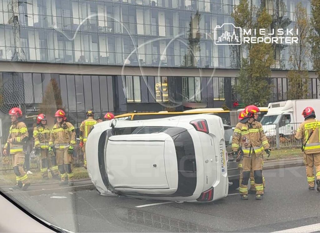 Grupa strażaków w mundurach i kaskach pracuje przy przewróconym samochodzie na ulicy obok nowoczesnego biurowca.