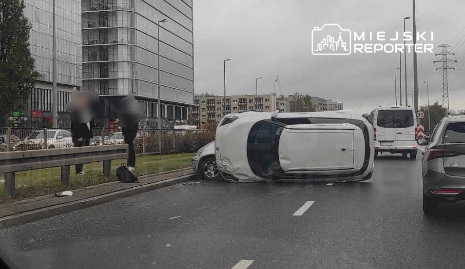 Samochód leżący na boku na jezdni, obok dwóch osób stojących przy barierce ochronnej, w tle nowoczesne budynki.
