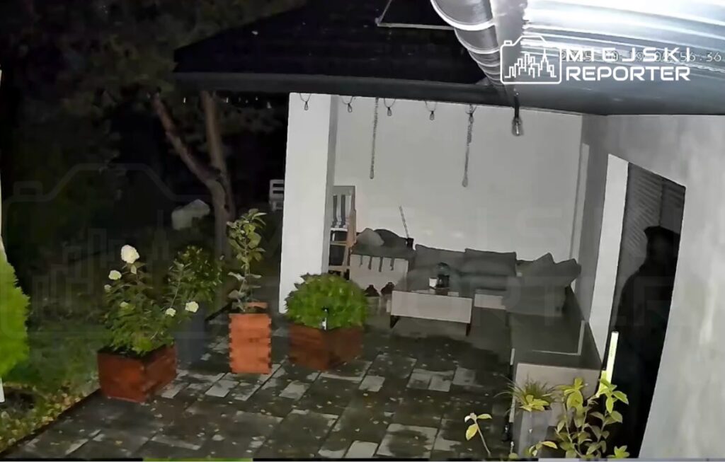 Oświetlony taras z meblami ogrodowymi, donicami z roślinami i płytkami na podłodze, otoczony ciemnym ogrodem.