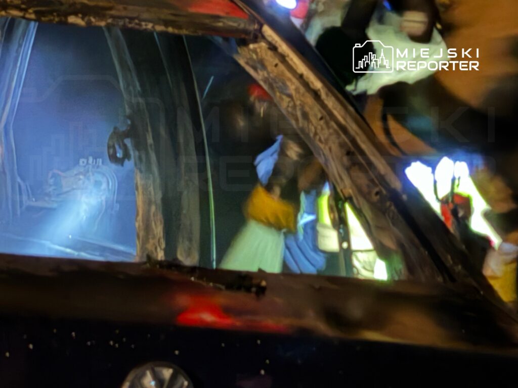 Ratownicy w odzieży ochronnej usuwają zniszczoną część pojazdu po wypadku, w tle widać niebieskie światło.